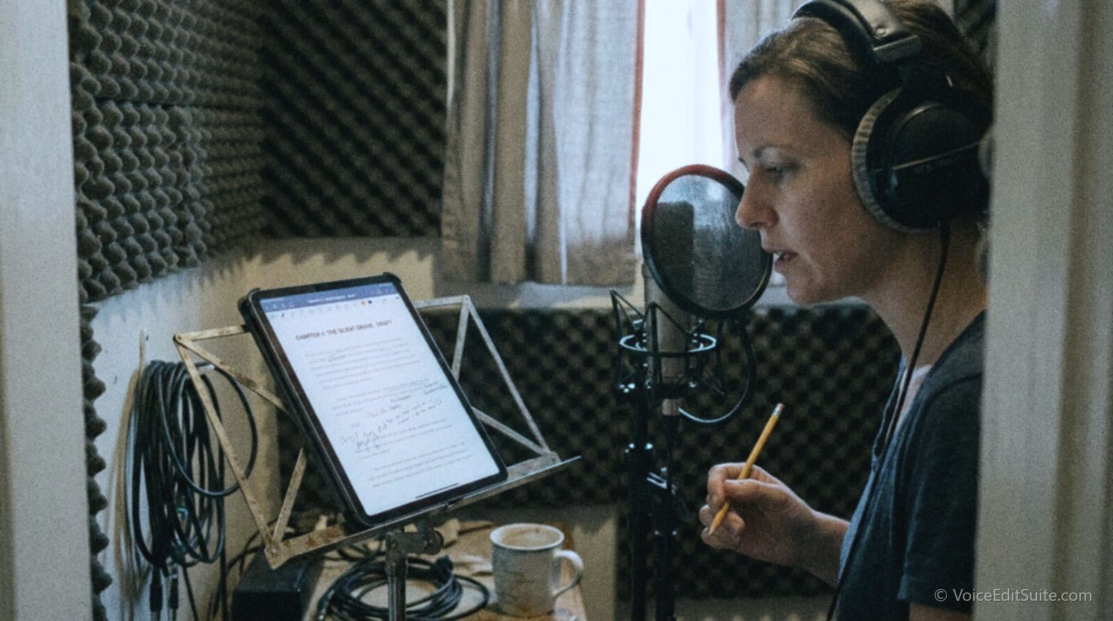 A narrator in a small foam-lined home booth recording a fantasy novel chapter, the marked-up manuscript on a tablet stand, headphones on, a pencil in hand