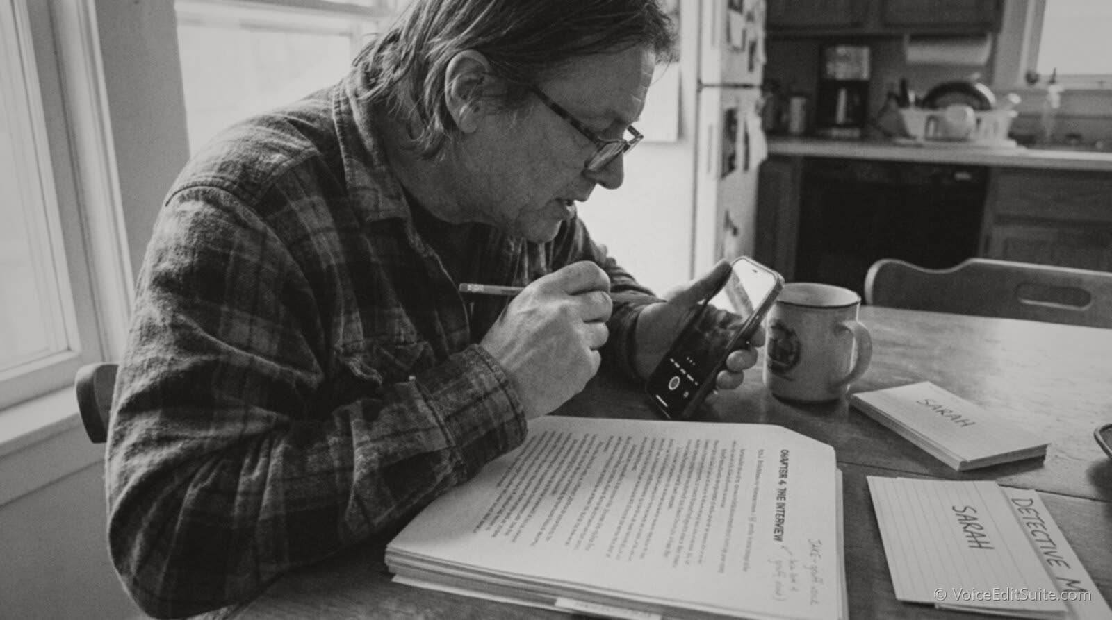 A narrator at a kitchen table during a prep read, a printed manuscript marked with coloured highlighters for each speaking character, a row of handwritten index cards with character names and a phone recording a voice note