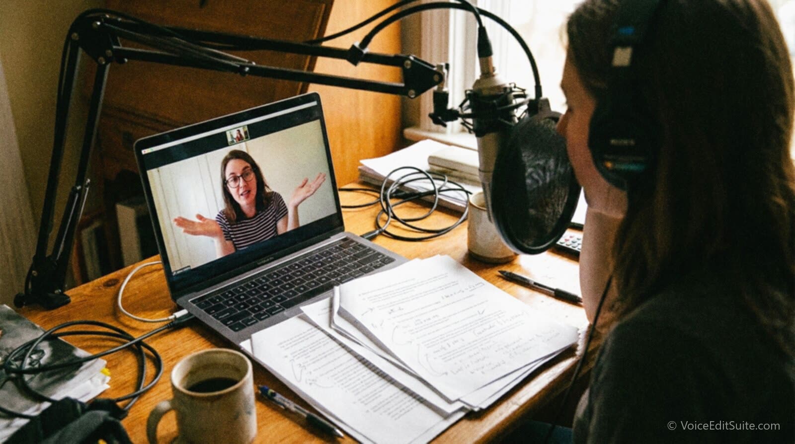 A voice actor at a laptop on a video call with a coach, a marked-up printed script on the desk, the actor leaning toward a microphone mid-sentence