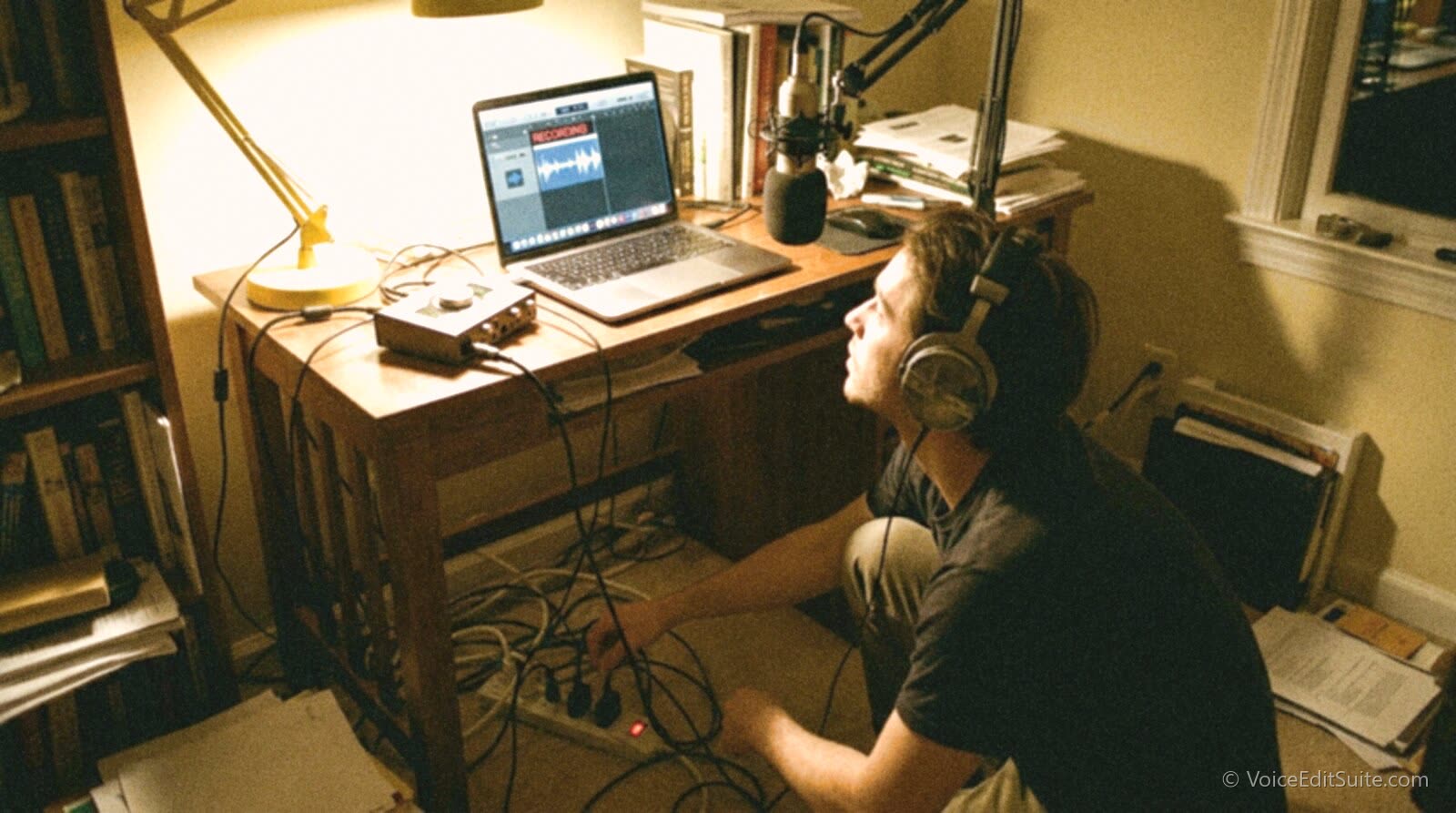 A podcaster in headphones unplugging a laptop charger from a power strip under a desk while watching the recording level meter on the laptop screen