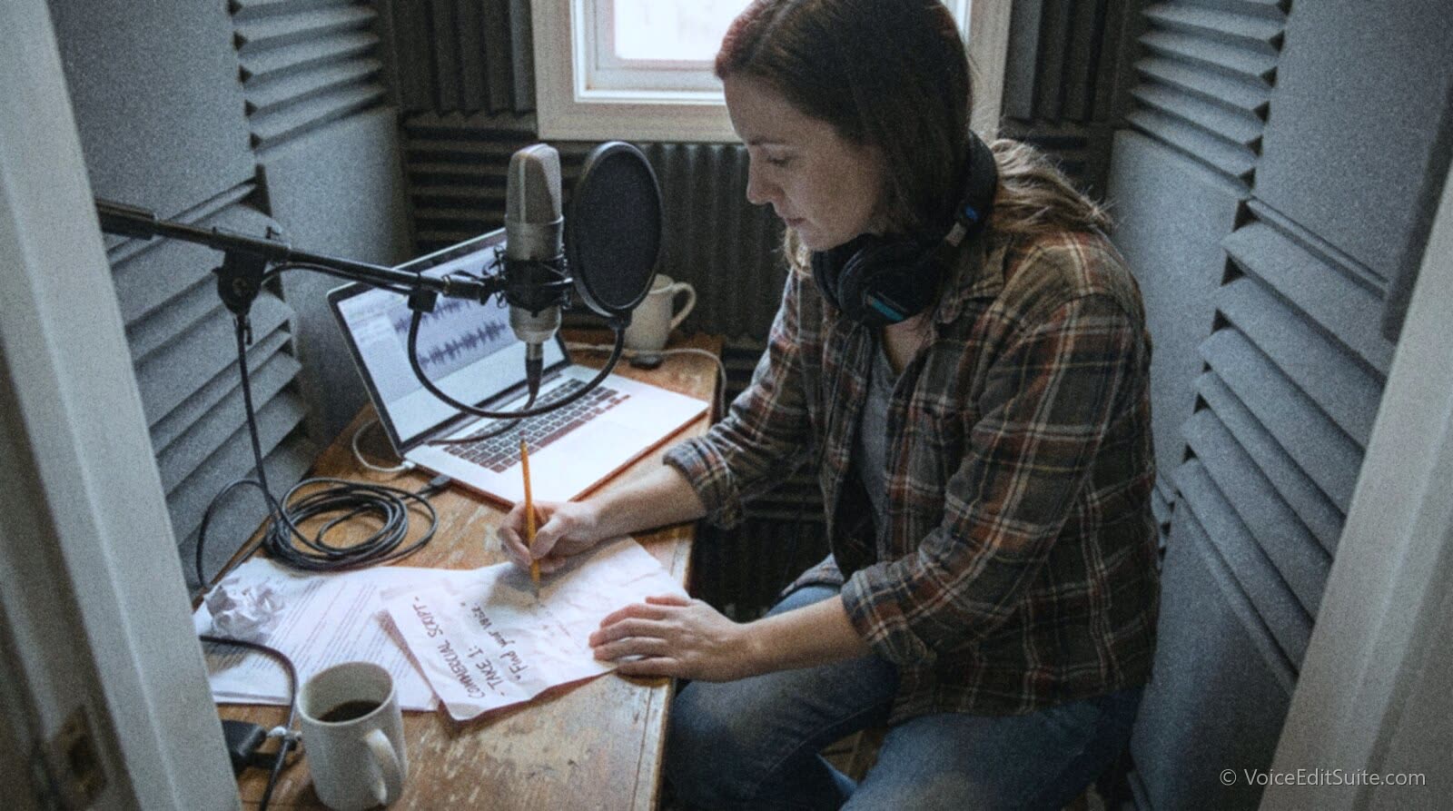 A voice actor at a desk in a small home booth writing the slate wording in pencil at the top of a printed commercial script, a microphone in front of her and a laptop recording screen beside it