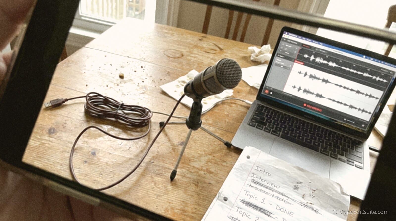 A phone snapshot of a USB microphone, a laptop running recording software and a handwritten run sheet on a kitchen table during a first podcast recording session
