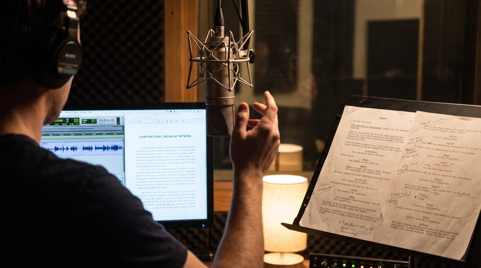 An audiobook narration session in progress: a microphone in a shock mount, a marked-up script on a stand, and a screen showing an audio waveform