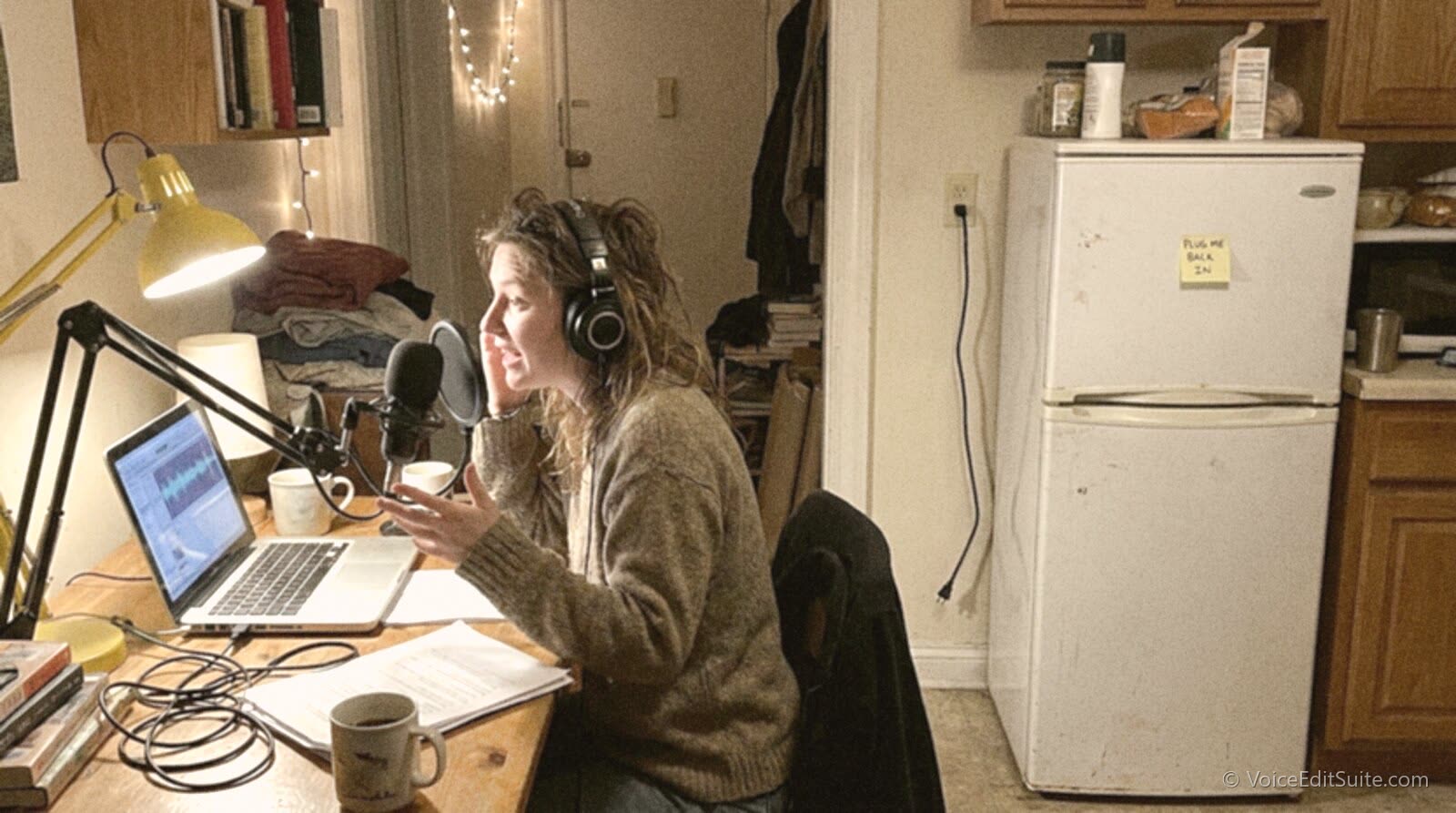 A podcaster at a desk in a small apartment recording with the fridge unplugged behind her and a sticky note on its door reading Plug me back in