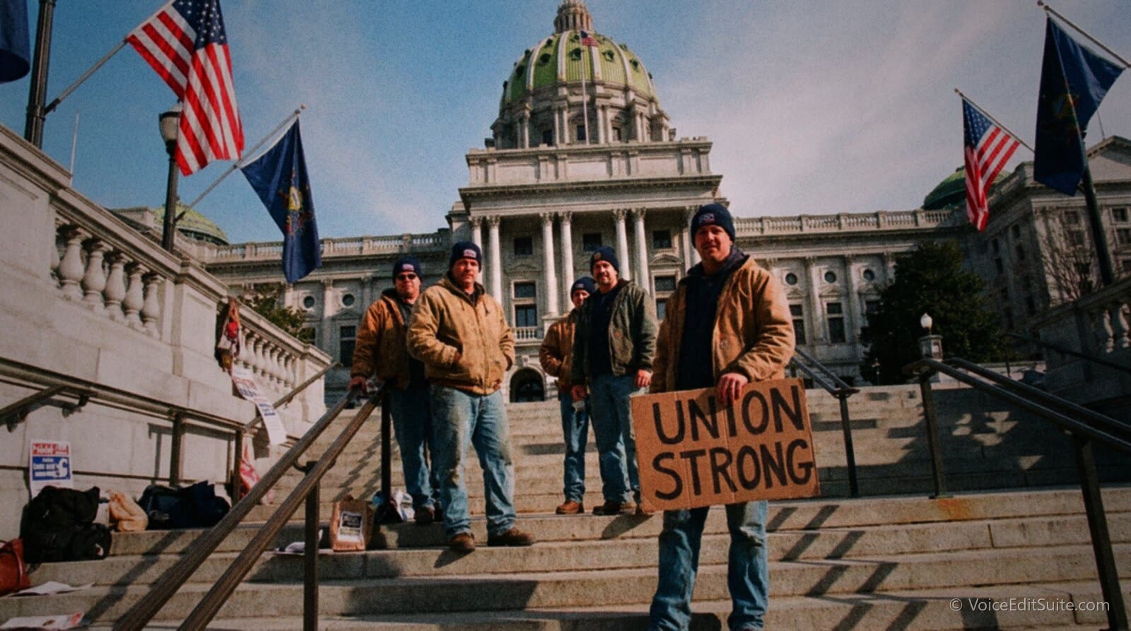 A state capitol building on a bright day with a union member holding a placard reading Union Strong on the steps