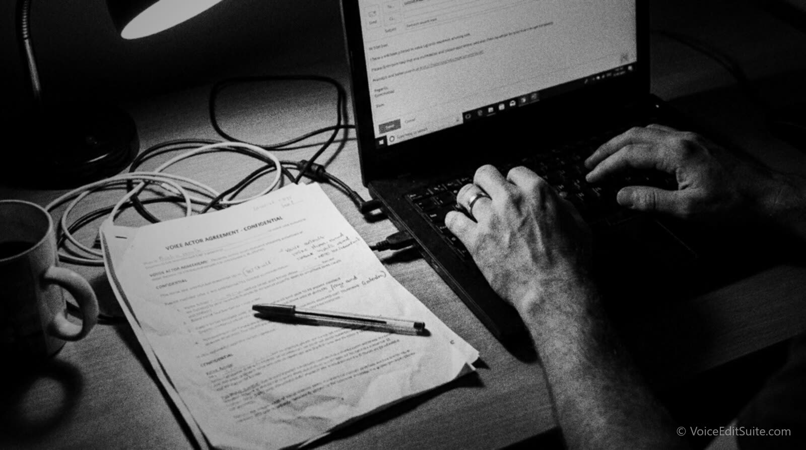 Black and white photo of a voice actor's hands typing an email on a laptop at a cluttered desk, with a printed union agreement document and a pen resting beside the keyboard