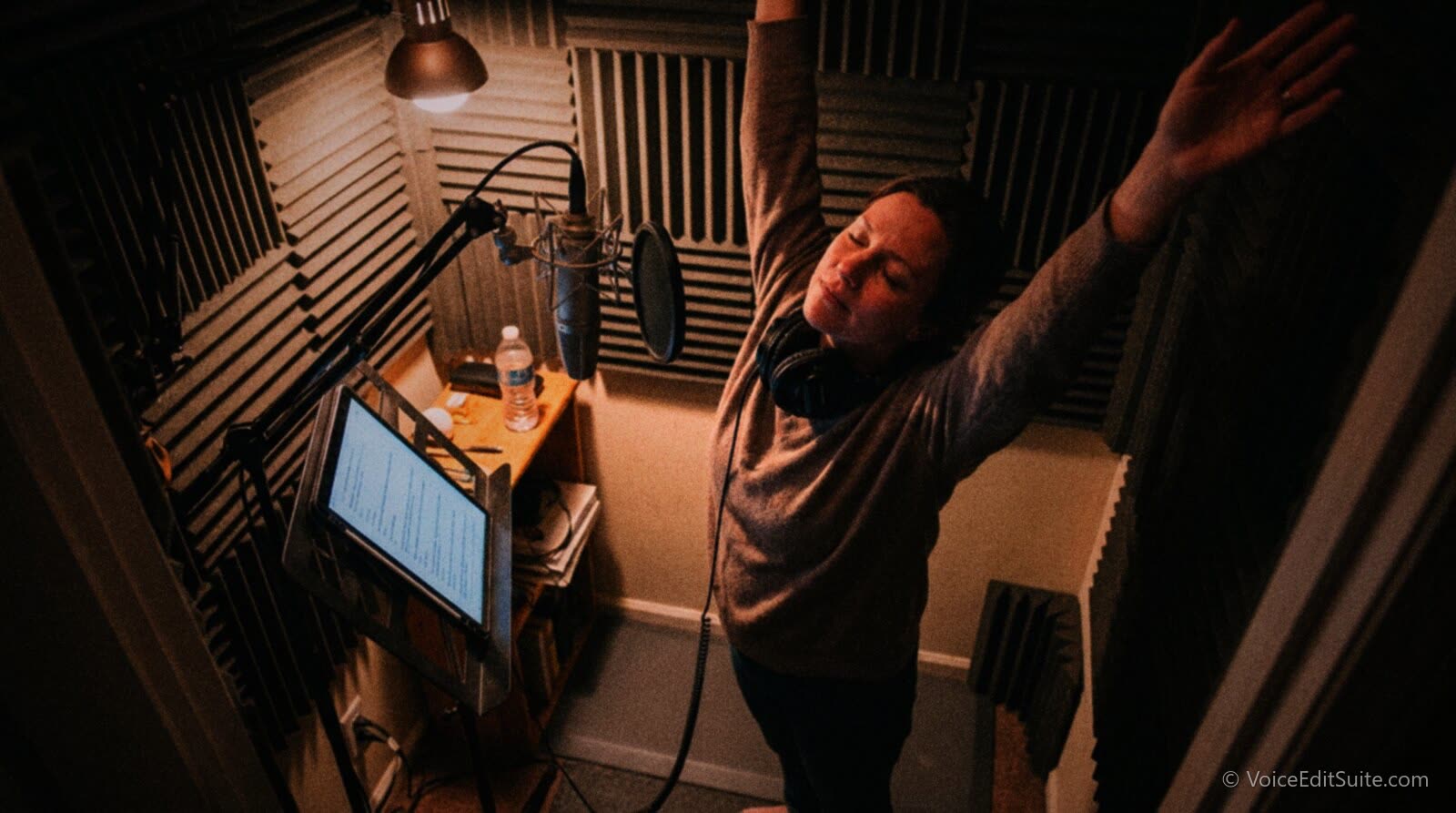 A narrator in a small home vocal booth stretching both arms overhead between takes, headphones around her neck, a tablet script on a stand at eye level and a microphone on a boom arm at mouth height beside her