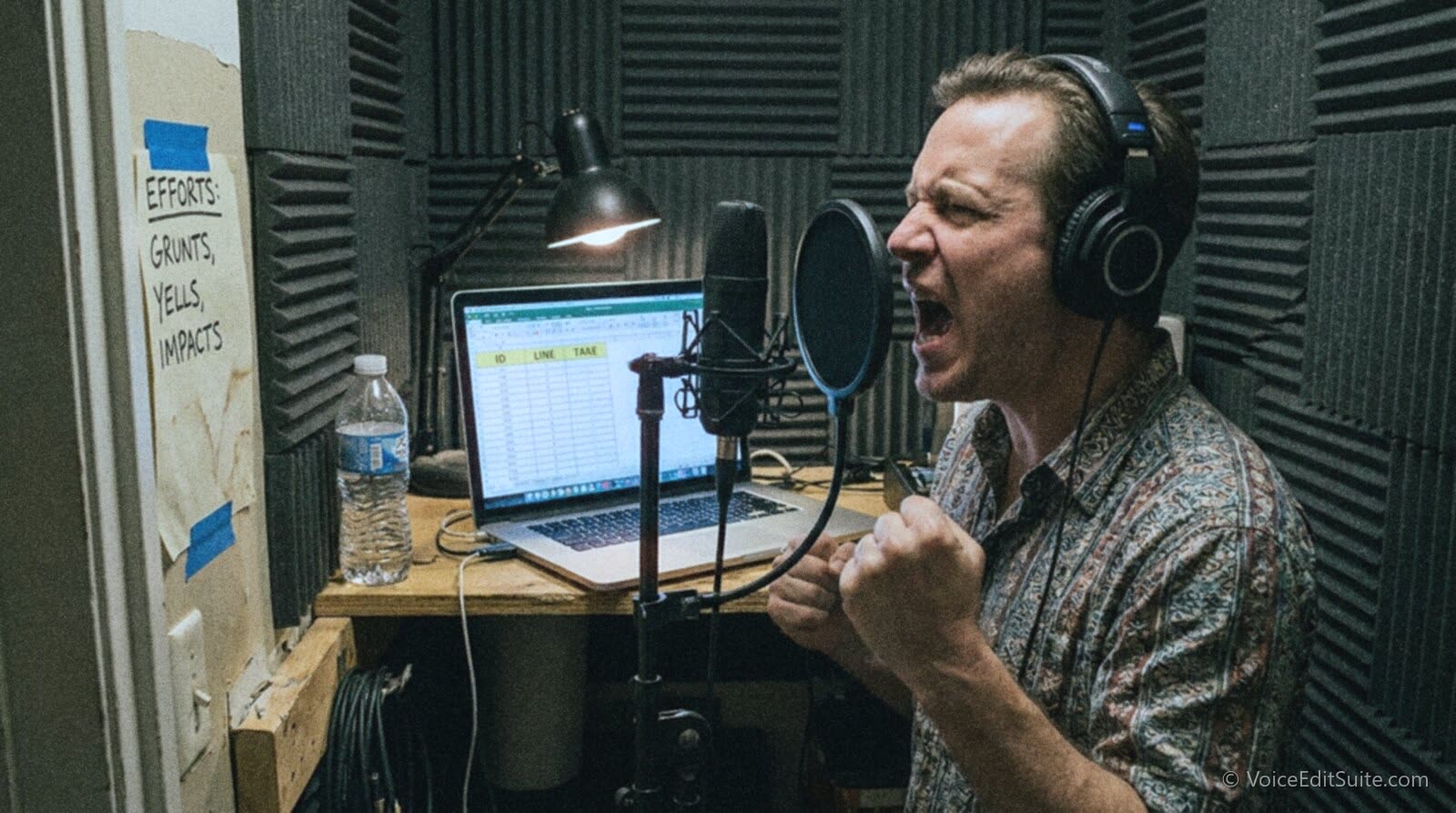 A voice actor in a small home vocal booth recording combat efforts for an indie game, a laptop beside the microphone showing a spreadsheet of numbered line IDs