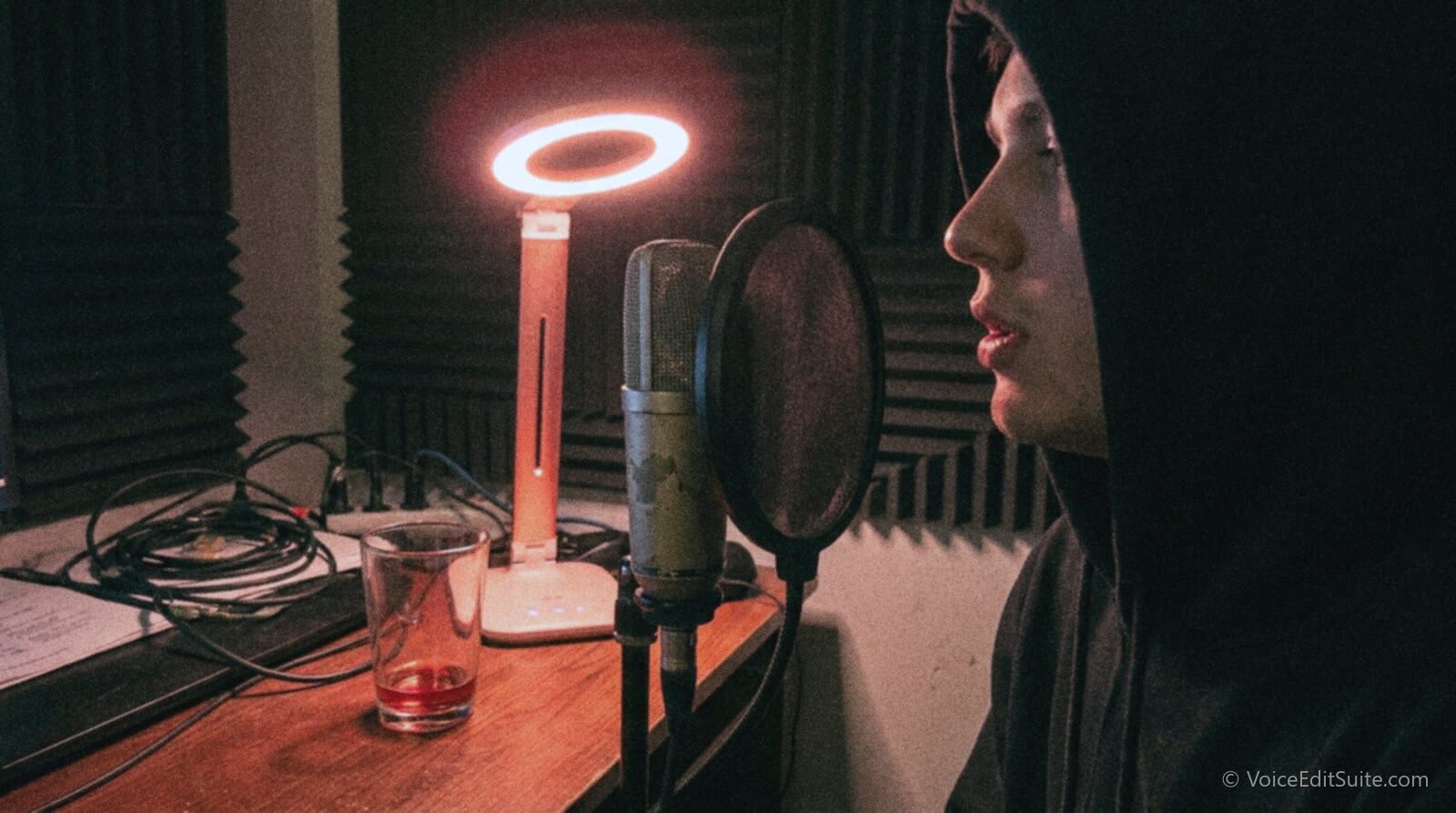 A narrator at a home mic stand at night, lit by a desk lamp with a red glow around a small LED, mid-way through a lip trill warm-up before recording